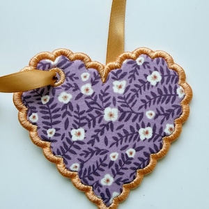 May include: A heart-shaped fabric ornament featuring a purple background with a repeating pattern of dark purple leaves and small white flowers. The edge is finished with gold embroidery and a gold ribbon loop is attached for hanging purposes.