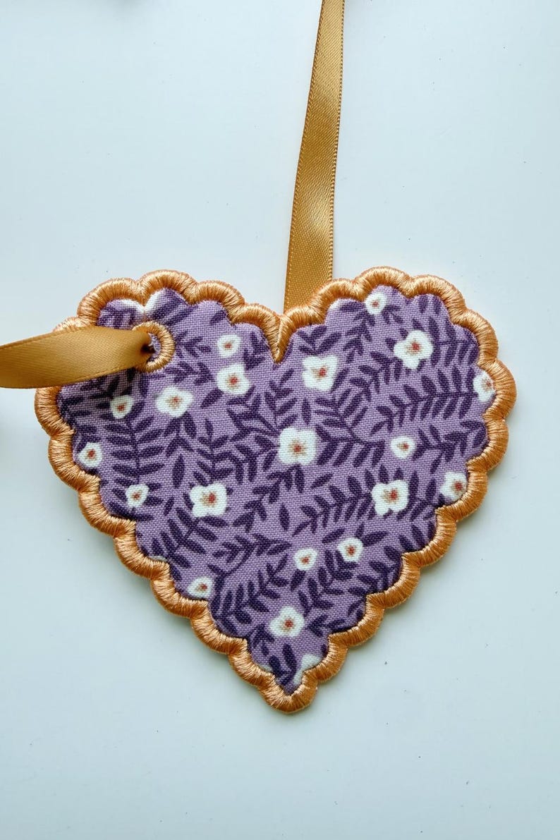 May include: A heart-shaped fabric ornament featuring a purple background with a repeating pattern of dark purple leaves and small white flowers. The edge is finished with gold embroidery and a gold ribbon loop is attached for hanging purposes.