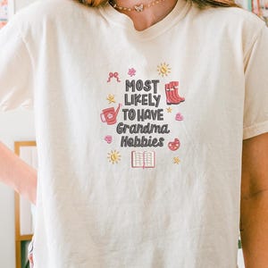 May include: Cream-colored t-shirt with the text "MOST LIKELY TO HAVE Grandma Hobbies" in a playful font. The design includes illustrations of a watering can, boots, a book, and other hobby-related icons. The shirt is a casual, comfortable style.