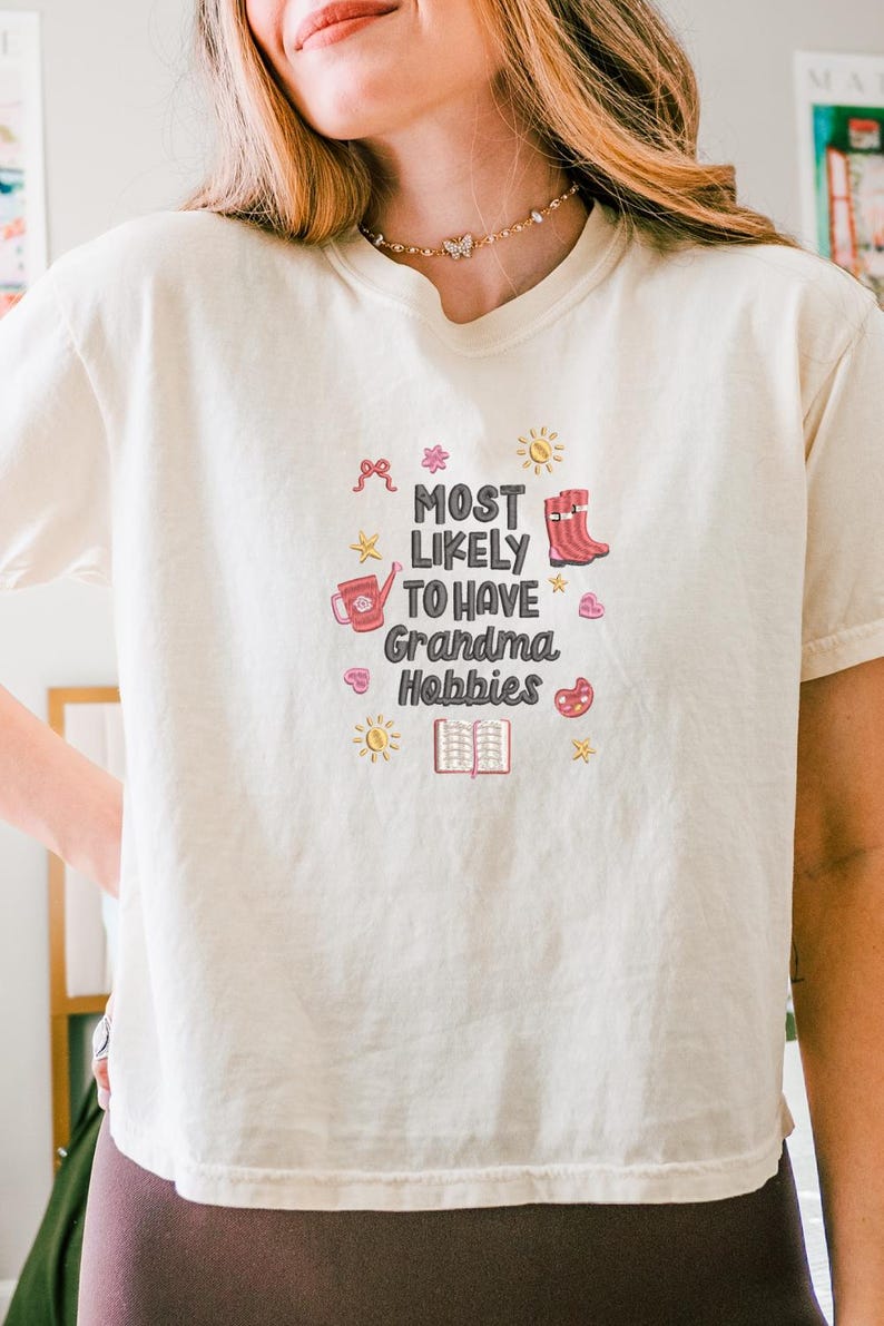 May include: Cream-colored t-shirt with the text "MOST LIKELY TO HAVE Grandma Hobbies" in a playful font. The design includes illustrations of a watering can, boots, a book, and other hobby-related icons. The shirt is a casual, comfortable style.
