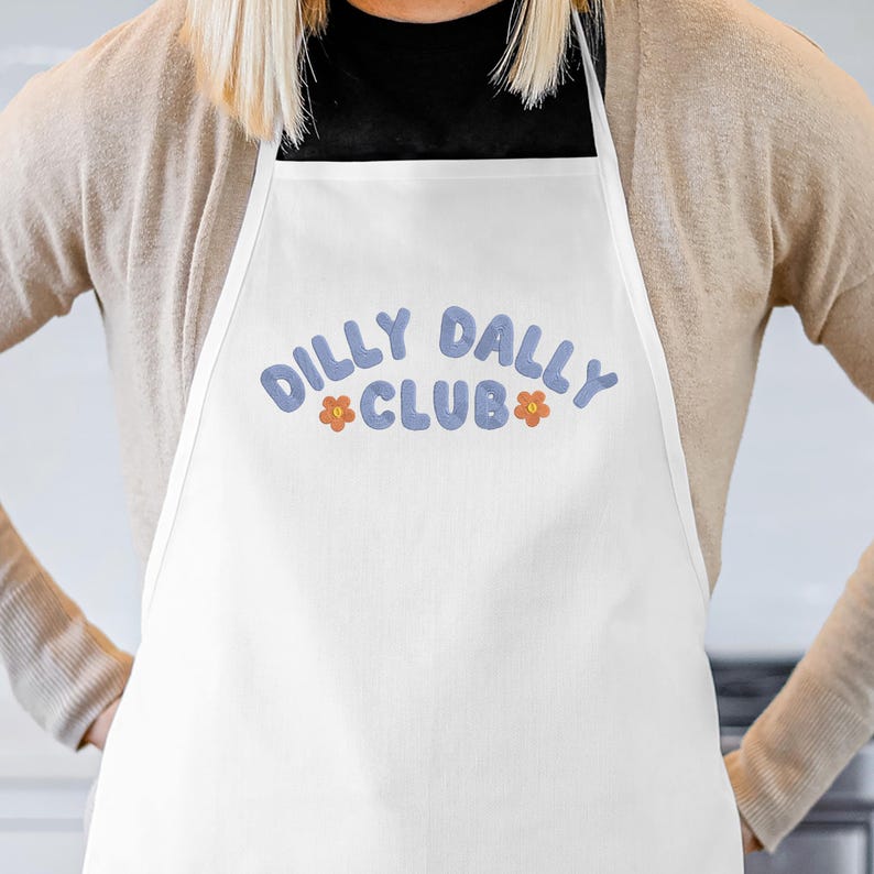 May include: White apron with the text "DILLY DALLY CLUB" in blue bubble letters, with orange flower accents. The apron is worn by a person wearing a tan cardigan and a black shirt.