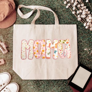 May include: A beige tote bag with the word "MAMA" in a colorful, floral design. The letters are filled with patterns of flowers, dots, and other designs. The bag has a simple, sturdy design with two handles.