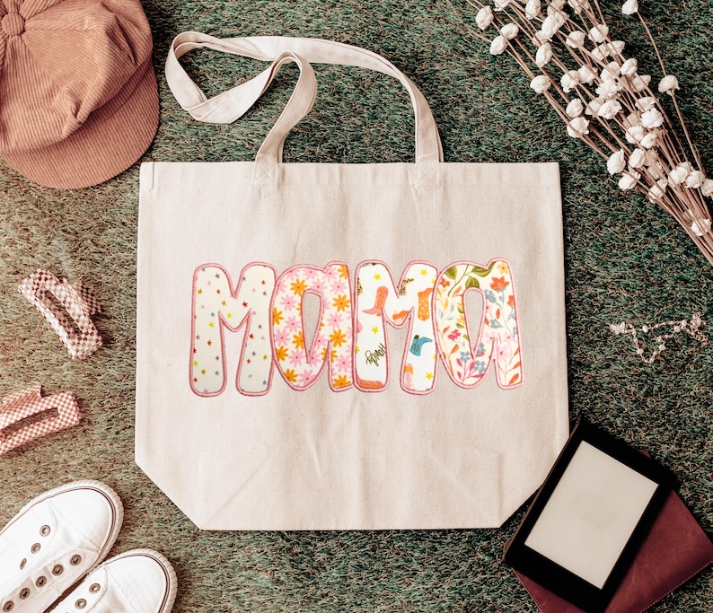 May include: A beige tote bag with the word "MAMA" in a colorful, floral design. The letters are filled with patterns of flowers, dots, and other designs. The bag has a simple, sturdy design with two handles.