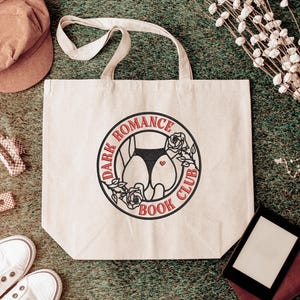 Op de afbeelding: Beige draagtas met de tekst "DARK ROMANCE BOOK CLUB" in een cirkelvormig ontwerp. Het ontwerp toont een silhouet van het onderlichaam van een persoon met een hart. De tas heeft twee handvatten en ligt op een groene ondergrond.