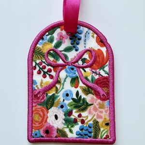 May include: A rectangular fabric tag with rounded top corners, featuring a vibrant floral pattern with red, orange, yellow, blue, and green flowers on a white background. The tag is finished with a bright pink stitched border and a matching ribbon.
