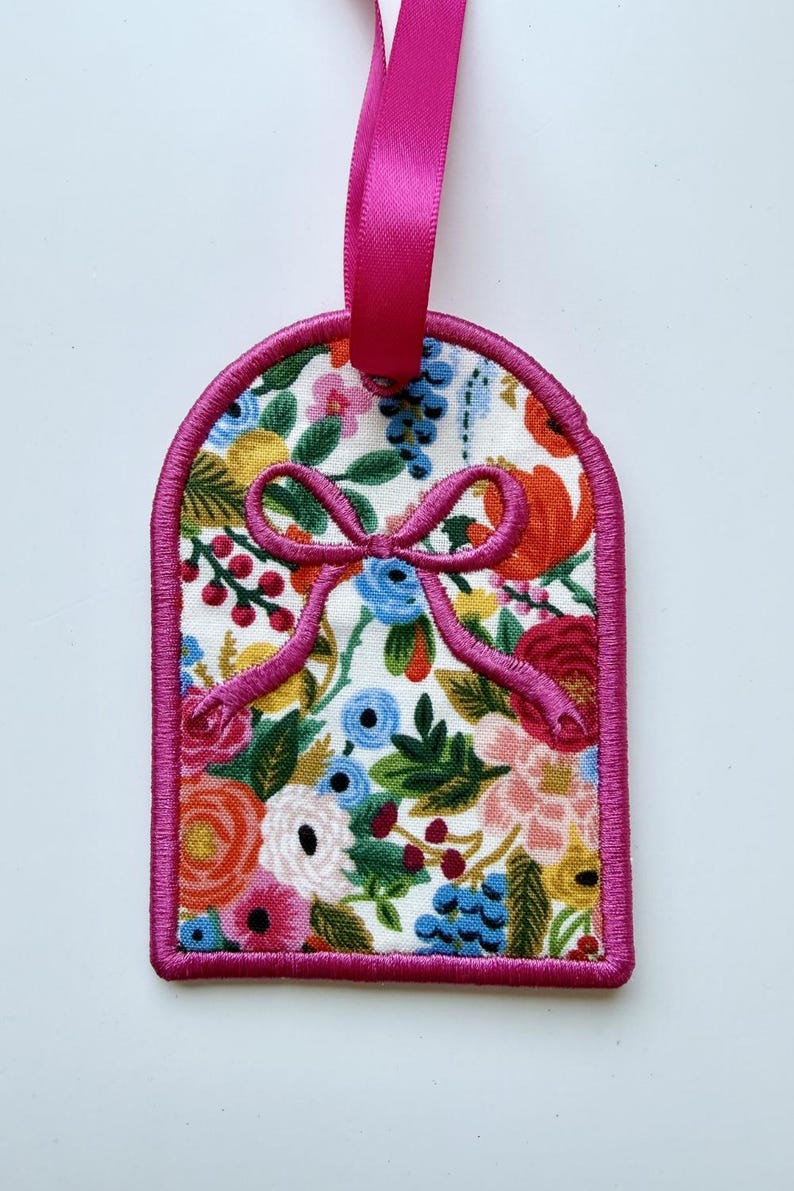 May include: A rectangular fabric tag with rounded top corners, featuring a vibrant floral pattern with red, orange, yellow, blue, and green flowers on a white background. The tag is finished with a bright pink stitched border and a matching ribbon.