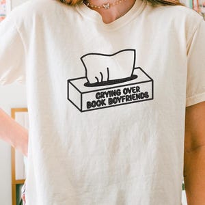 Puede incluir: Camiseta color crema con el texto "Crying Over Book Boyfriends" en negro. El gr&aacute;fico presenta una caja de pa&ntilde;uelos con pa&ntilde;uelos saliendo. La camiseta es de estilo casual y de manga corta.