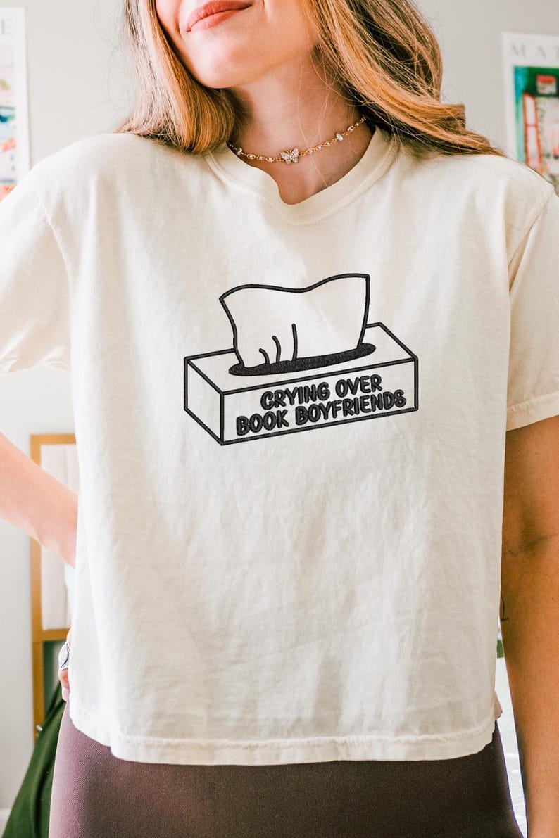 Puede incluir: Camiseta color crema con el texto "Crying Over Book Boyfriends" en negro. El gr&aacute;fico presenta una caja de pa&ntilde;uelos con pa&ntilde;uelos saliendo. La camiseta es de estilo casual y de manga corta.