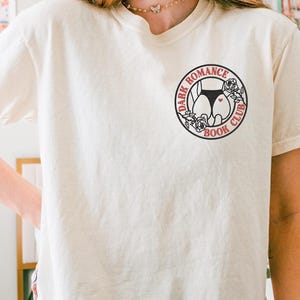 Op de afbeelding: Cr&egrave;mekleurig T-shirt met een cirkelvormige afbeelding met de tekst "DARK ROMANCE BOOK CLUB" rond een ontwerp van een silhouet van een onderlichaam in zwart en rood.