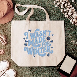 May include: Beige tote bag with the phrase "I WASN'T MADE FOR WINTER" in blue script. The bag is surrounded by a brown corduroy cap, white sneakers, and other accessories on a green surface.