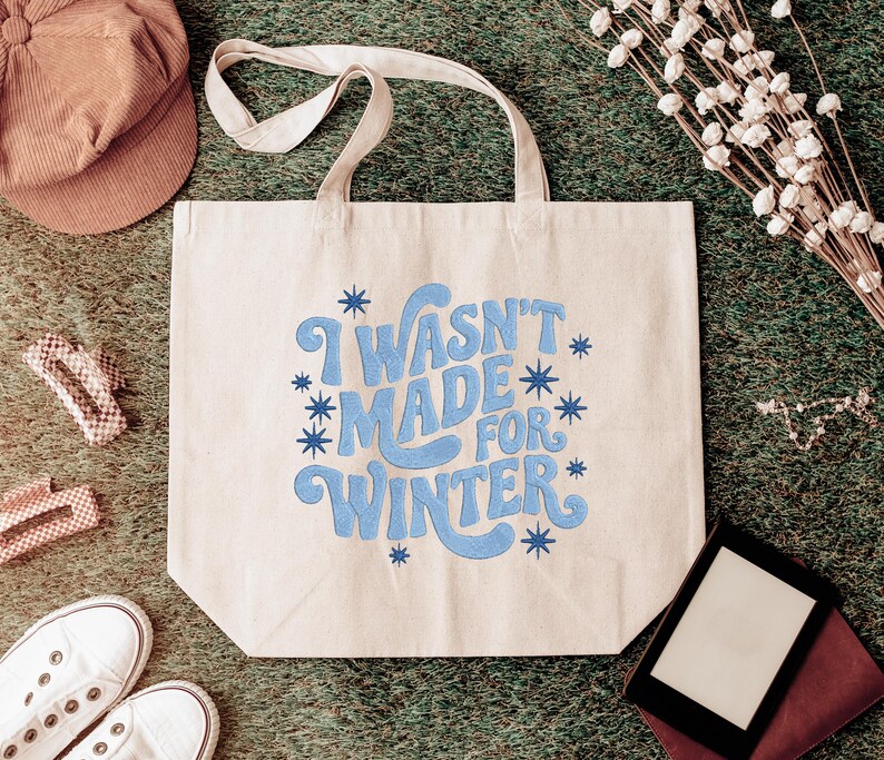 May include: Beige tote bag with the phrase "I WASN'T MADE FOR WINTER" in blue script. The bag is surrounded by a brown corduroy cap, white sneakers, and other accessories on a green surface.