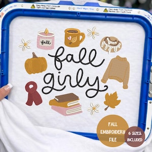 May include: An embroidery design on white fabric featuring the text fall girly in black script. Surrounding the text are embroidered icons including a pink candle, a brown mug, white flowers, a pumpkin, a sweater, a scarf, books, and a leaf.