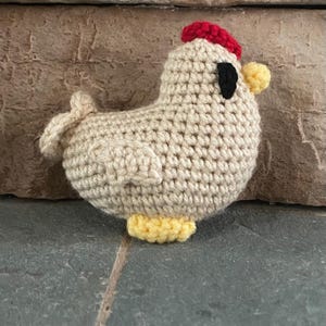 May include: A small, beige crochet chicken figurine featuring a bright red comb on top of its head, a yellow beak, yellow feet, and a single black eye. The body is crafted with a textured yarn stitch pattern, showing a rounded shape with a small tail.