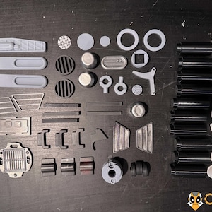 May include: An assortment of grey and black 3D-printed mechanical components, including cylinders, circular parts, and rectangular pieces. The text "KIT 001" is visible. The Creative Mechanics logo is in the bottom right corner.