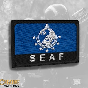 May include: A rectangular embroidered patch featuring a blue background with a white globe emblem surrounded by stars and the white text SEAF on a black horizontal stripe at the bottom. The patch has a black border and a textured fabric finish.