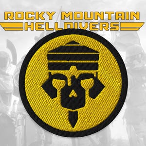 Pode incluir: Patch bordado com o texto "ROCKY MOUNTAIN HELLDIVERS" em amarelo. O patch apresenta um design circular preto e amarelo com um emblema de caveira estilizado. O fundo é uma imagem desfocada de figuras em armadura futurista.