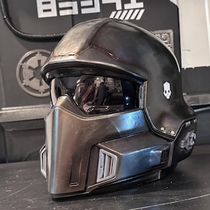 May include: A dark metallic gray full-face helmet featuring a prominent angular visor, a central nose guard, and side vents. The helmet displays a small white skull emblem on the side and has a smooth, weathered finish with visible scuffs.