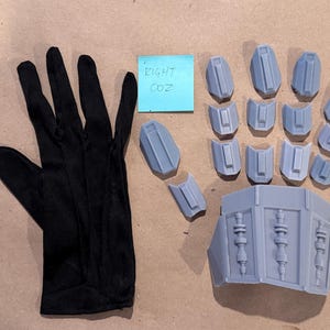 Könnte beinhalten: Ein schwarzer Stoffhandschuh neben einem Set aus hellgrauen, starren, geometrischen Rüstungsteilen für Hand und Unterarm. Ein kleiner blauer quadratischer Zettel mit dem handschriftlichen Text 'EIGHT 002' liegt zwischen dem Handschuh und den Teilen.