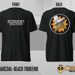 May include: A charcoal black triblend t-shirt with a white graphic on the front that says "L771A3M7" and "77007 707MT7". The back of the shirt features a white boot stomping on a brown spaceship with a yellow starburst behind it.
