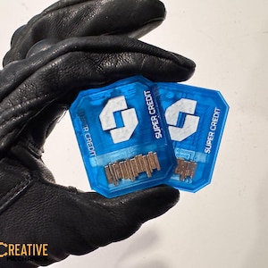 Puede incluir: Dos tarjetas Super Credit azules con logotipos y texto blancos se sostienen en un guante de cuero negro. Las tarjetas tienen forma rectangular con esquinas redondeadas y una serie de elementos metálicos marrones.