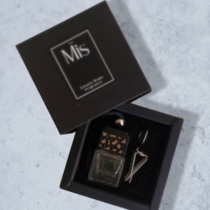 May include: A black box with the word "Mis" and "Luxury Home Fragrance" printed on it. The box contains a small black box with a black and gold car air freshener inside.
