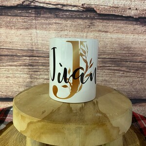 Peut inclure: Une tasse en céramique blanche avec une lettre dorée "J" et un motif floral. La tasse est sur un support en bois.