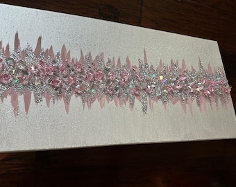 Pode incluir: Uma pintura em tela branca com uma faixa horizontal de glitter rosa e prateado. O glitter está disposto em um padrão de onda e inclui pequenos cristalls rosa e iridescentes.