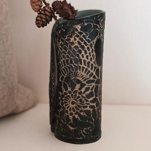 May include: A dark blue ceramic vase with a lace-like pattern, holding a branch with pine cones and a dark flower. The vase is cylindrical and sits on a white surface. A beige cushion is in the background.