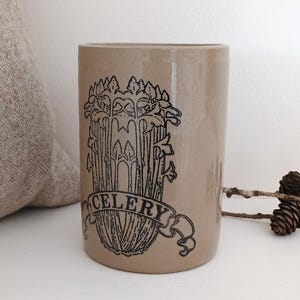 May include: A beige ceramic cylinder vase featuring a black illustration of celery and the word "CELERY" on a banner. The vase is on a white surface, with a beige cushion and a branch with pine cones in the background.