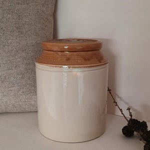 May include: A cream-coloured stoneware jar with a brown rim and lid. The cylindrical jar is displayed on a white surface. A decorative pillow and a sprig of pinecones are in the background.