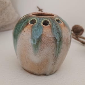 May include: A ceramic bud vase with a rounded form and three openings. The vase features a mottled glaze in brown, green, and blue hues. It is designed to hold a few flower stems. The overall aesthetic is rustic and handmade, with a unique, organic feel.