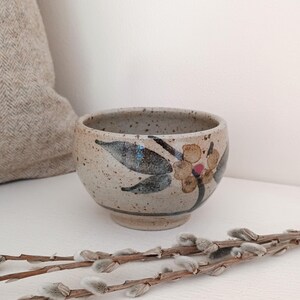 May include: A small, round ceramic bowl with a speckled beige glaze. The exterior features a hand-painted floral design with dark green leaves, brown stems, and a single pink centre dot on the flower. The bowl has a short, raised foot base.