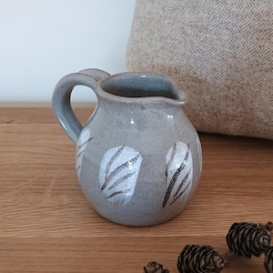 May include: A small, grey ceramic jug with a rounded body and a curved handle. The jug features white and black feather-like designs. The jug is sitting on a wooden surface.