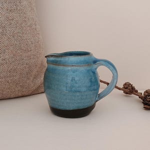 May include: A small ceramic jug featuring a rounded body with a glossy blue glaze that transitions into a dark brown base. The jug has a curved handle on the side and a small pouring spout at the top rim, showcasing a handcrafted aesthetic.