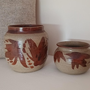 May include: Two ceramic pots with a speckled beige finish and brown painted floral designs. The larger pot is cylindrical, and the smaller one is round. Both have a brown rim.