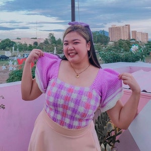 Puede incluir: Una mujer lleva una blusa de crochet morado y blanco con escote cuadrado y mangas abullonadas. La blusa tiene un patr&oacute;n de cuadros. Lleva una falda rosa.