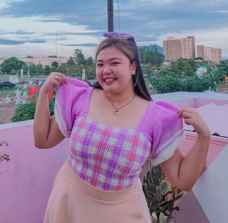Puede incluir: Una mujer lleva una blusa de crochet morado y blanco con escote cuadrado y mangas abullonadas. La blusa tiene un patr&oacute;n de cuadros. Lleva una falda rosa.