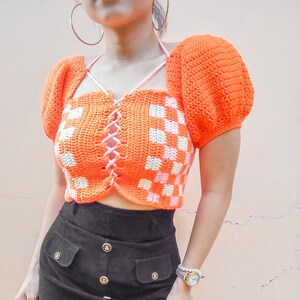 Puede incluir: Un top corto de crochet con un patr&oacute;n de cuadros en naranja y blanco. El top tiene un frente con cordones y mangas abullonadas.