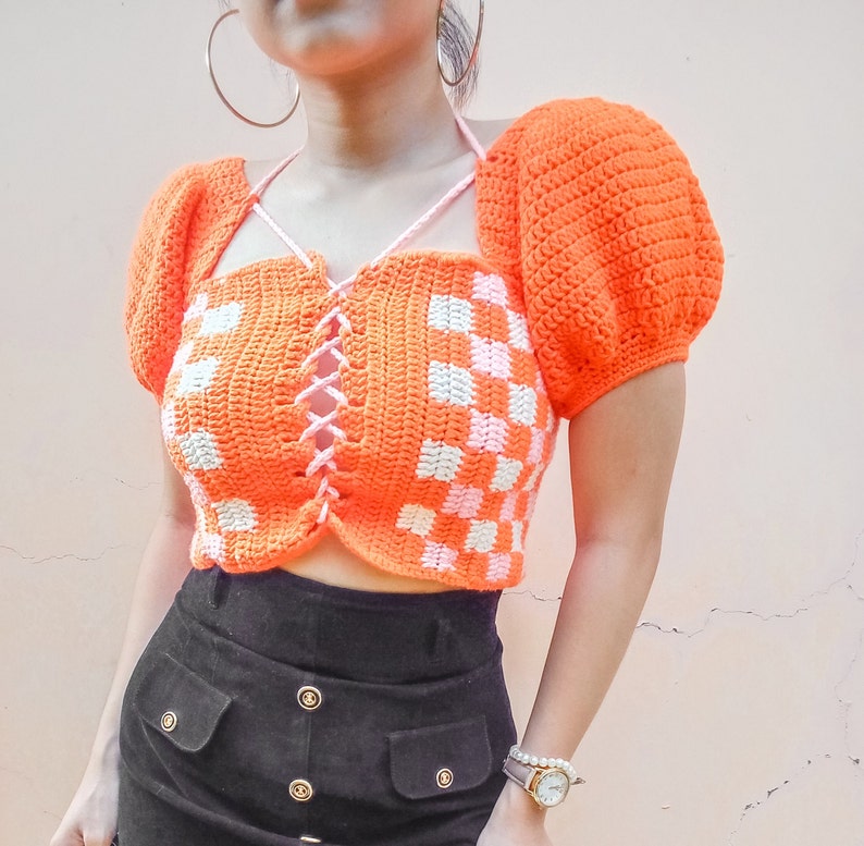Puede incluir: Un top corto de crochet con un patr&oacute;n de cuadros en naranja y blanco. El top tiene un frente con cordones y mangas abullonadas.