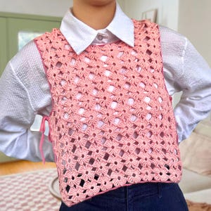 Könnte beinhalten: Hellrosa gehäkelte Weste mit floralem Muster, getragen über einem weißen Hemd mit Kragen und dunkelblauen Jeans. Die Weste hat rosa Bänder an der Seite.