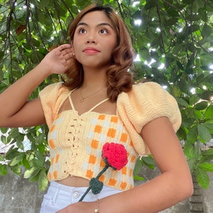 Puede incluir: Un top de crochet de cuadros amarillos y blancos con mangas abullonadas y un frente con cordones. La modelo sostiene una rosa roja de crochet.