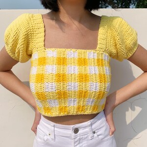 Puede incluir: Un top de crochet de cuadros amarillos y blancos con mangas abullonadas. El top es corto y tiene un escote cuadrado.