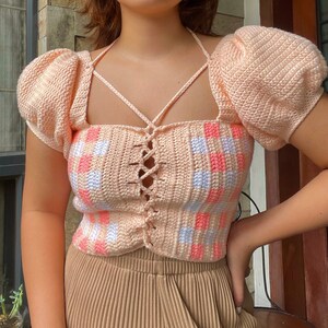 Puede incluir: Un top de crochet rosa con un patr&oacute;n cuadrado y un cord&oacute;n en la parte delantera. El top tiene mangas abullonadas y un escote cuadrado.