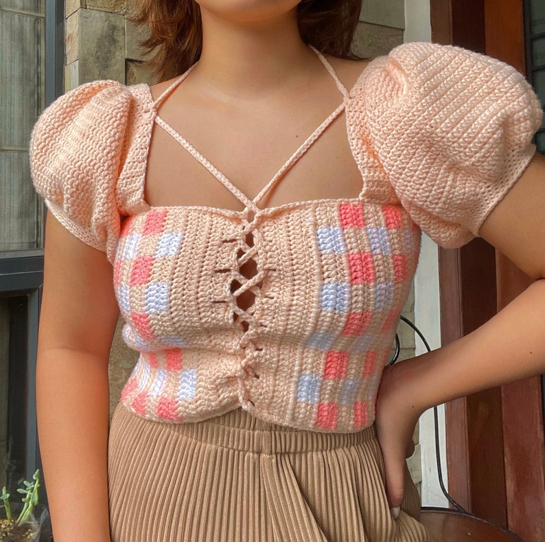 Puede incluir: Un top de crochet rosa con un patr&oacute;n cuadrado y un cord&oacute;n en la parte delantera. El top tiene mangas abullonadas y un escote cuadrado.