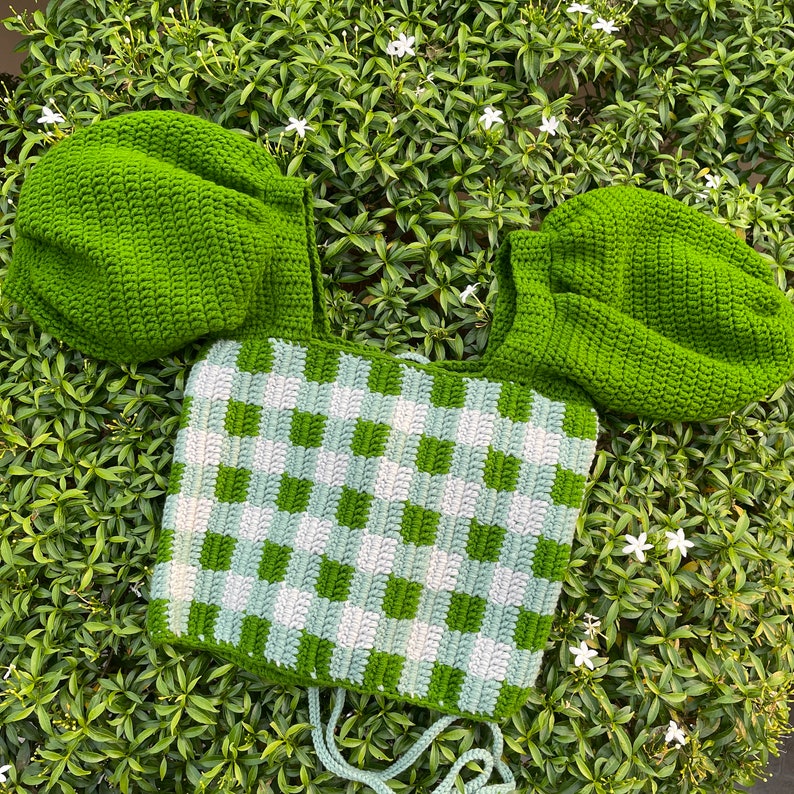 Puede incluir: Top de crochet verde con un patr&oacute;n de cuadros y mangas abullonadas. El top est&aacute; colocado sobre una cama de follaje verde.