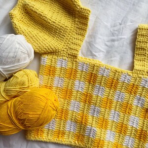 Puede incluir: Un top de crochet amarillo y blanco con un patr&oacute;n de cuadros. El top est&aacute; hecho con un escote cuadrado y tiene un gorro de crochet en la parte superior.