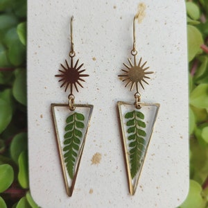 May include: Gold earrings with a triangle-shaped pendant featuring a pressed green fern leaf. The top of the earring has a gold starburst design.