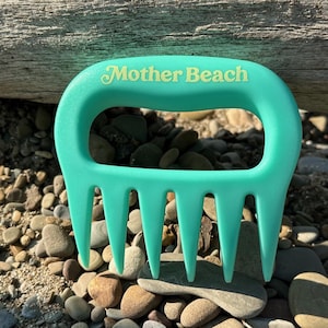 May include: A turquoise beach comb with the words "Mother Beach" printed on the handle. The comb is resting on a bed of pebbles and small stones, with a weathered log in the background.