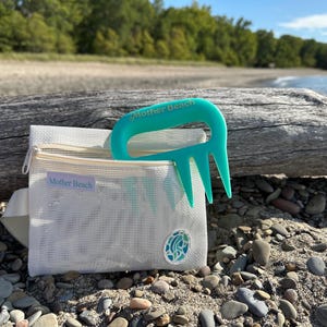 May include: A white mesh bag with a zipper, containing a teal beach rake. The bag has a label that says "Mother Beach". The rake is a bright teal color and has a handle and tines. The bag is sitting on a sandy beach with rocks.