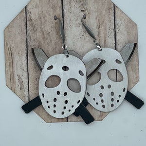 May include: A pair of silver earrings featuring a white hockey mask with black accents and a crossed hockey stick design.